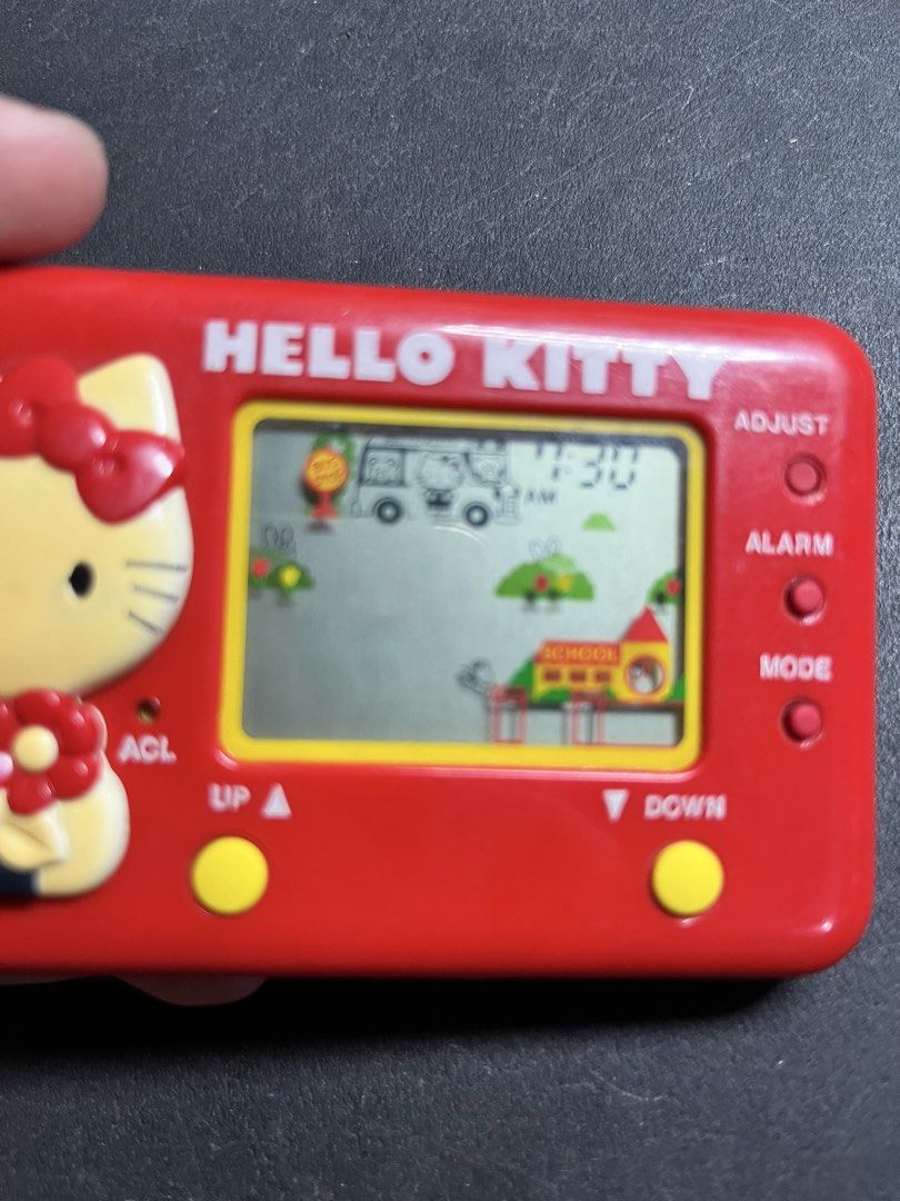 Vintage Hello Kitty Tomy LCD Game Electronic Handheld (Category: Interactive Toys) located in Caloocan City. Submitted by snapshop_ph (Listing ID 1471018923, Image 2). Description: Red handheld LCD game from Sanrio and Tomy. Kitty character molded on the left side holding a flower. Digital screen powers on, displays clock and game graphics like a school building and bus stop. Yellow control buttons below the screen. Battery cover on back, origina