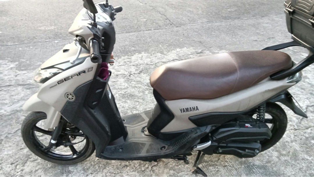 Yamaha Mio Gear S Scooter (Category: Motorbikes for Sale) located in Quezon City. Submitted by ElaineCastillo (Listing ID 1471052373, Image 2). Description: Yamaha Mio Gear S scooter in matte beige with a brown seat. Includes a rear storage box mounted on the back. Some minor scuffs and scratches visible on the front fairing and fender. Electric start, black alloy wheels, and blue accent graphics on the handlebar cowl. Clean floorboard and ready 