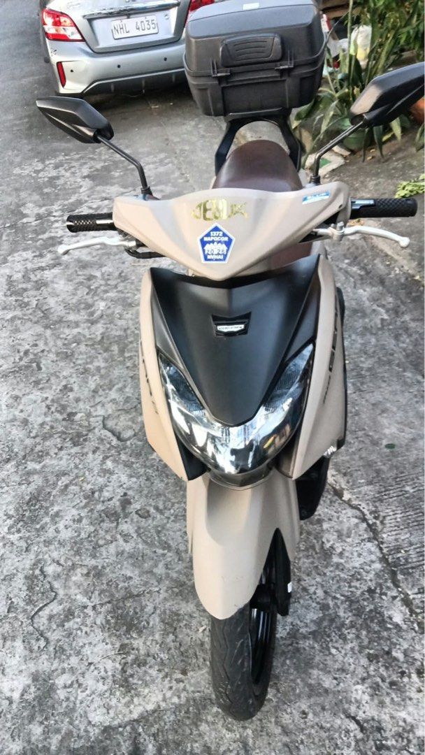 Yamaha Mio Gear S Scooter (Category: Motorbikes for Sale) located in Quezon City. Submitted by ElaineCastillo (Listing ID 1471052373, Image 1). Description: Yamaha Mio Gear S scooter in matte beige with a brown seat. Includes a rear storage box mounted on the back. Some minor scuffs and scratches visible on the front fairing and fender. Electric start, black alloy wheels, and blue accent graphics on the handlebar cowl. Clean floorboard and ready 