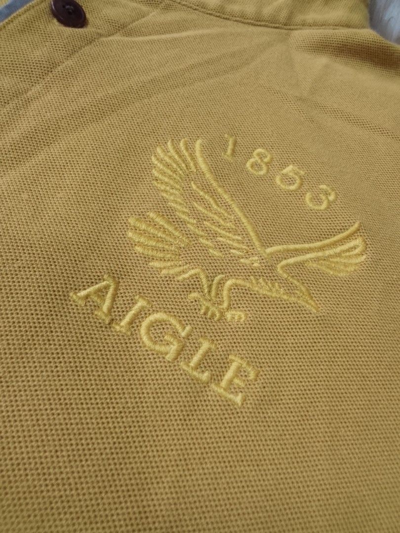 Aigle CT115 "EAGLETWIST" POLO SHIRT (Category: Tshirts & Polo Shirts ) located in Manila. Submitted by jctclthng_2026 (Listing ID 1465622683, Image 2). Description: - Brand: Aigle (Est. 1853) - Color: Mustard yellow with contrasting grey inner collar and placket - Material: Classic pique cotton fabric - Details: Three dark brown branded buttons, embroidered eagle logo on the left chest - Fit: Short sleeves, standard hem - Condition: Clean, well-m