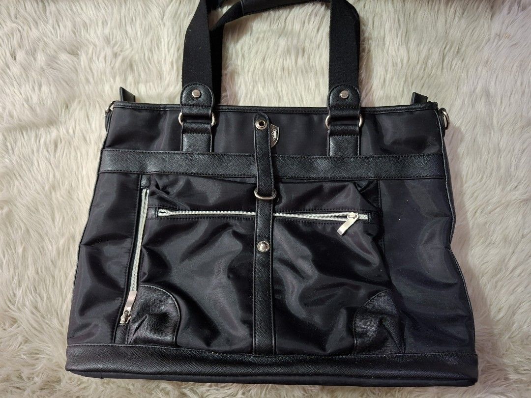 Black Nylon Tote Bag (Category: Tote Bags) located in Caloocan City. Submitted by bycara (Listing ID 1465642162, Image 1). Description: Black nylon tote bag with faux leather trim and silver tone hardware. Dual carry handles on top. Front features a vertical zipper pocket, a flap pocket, and a central strap snap closure. Back includes a large open pocket and side zipper expansion. Interior lined in brown fabric with an inner zipped compartment an