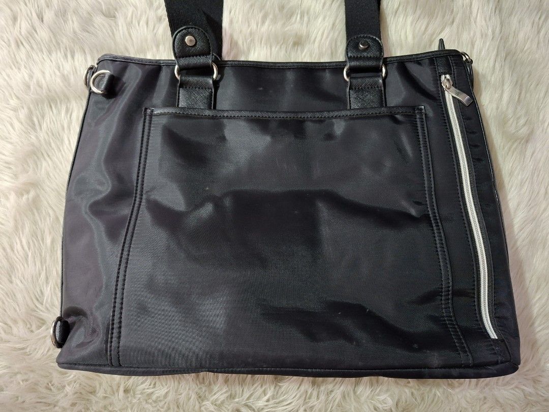 Black Nylon Tote Bag (Category: Tote Bags) located in Caloocan City. Submitted by bycara (Listing ID 1465642162, Image 2). Description: Black nylon tote bag with faux leather trim and silver tone hardware. Dual carry handles on top. Front features a vertical zipper pocket, a flap pocket, and a central strap snap closure. Back includes a large open pocket and side zipper expansion. Interior lined in brown fabric with an inner zipped compartment an