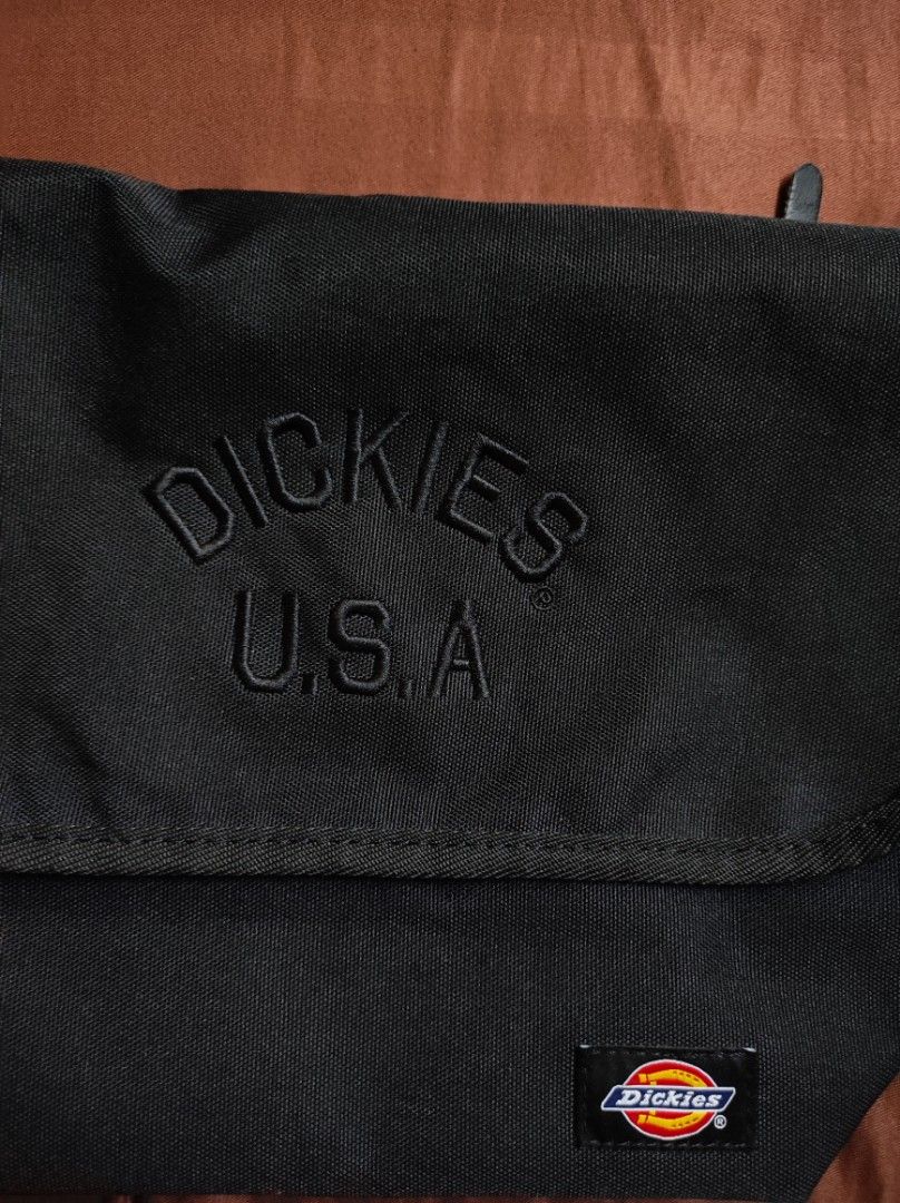 Dickies USA Black Messenger Bag (Category: Sling Bags) located in Batangas. Submitted by astrofinds_ (Listing ID 1465597709, Image 2). Description: - Brand: Dickies - Material: Durable black canvas - Design: Embossed Dickies USA text and classic woven logo patch on the front flap - Closure: Secure zip top under flap with snap button closure - Strap: Adjustable shoulder strap for comfortable wear - Interior: Branded lining with an extra zippered p