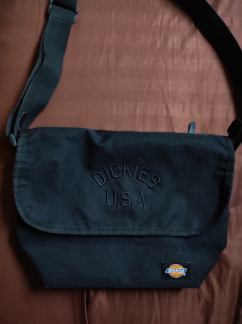 Dickies USA Black Messenger Bag (Category: Sling Bags) located in Batangas. Submitted by astrofinds_ (Listing ID 1465597709, Image 1). Description: - Brand: Dickies - Material: Durable black canvas - Design: Embossed Dickies USA text and classic woven logo patch on the front flap - Closure: Secure zip top under flap with snap button closure - Strap: Adjustable shoulder strap for comfortable wear - Interior: Branded lining with an extra zippered p