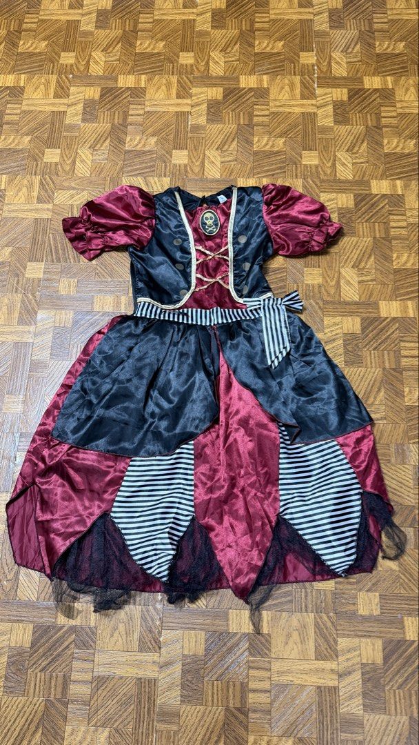 Pirate Dress Costume from US (Category: Babies & Kids Fashion) located in Quezon City. Submitted by frappes (Listing ID 1465702009, Image 1). Description: - Size: Kids aged 9-10 years (Height: 134-140cm) - Material: 100% polyester - Design: Black and red satin-style dress with gold skull patch, striped panels, and attached sash - Details: Black tulle underlay at the hem for extra volume - Perfect for: Halloween, school events, and dress-up partie