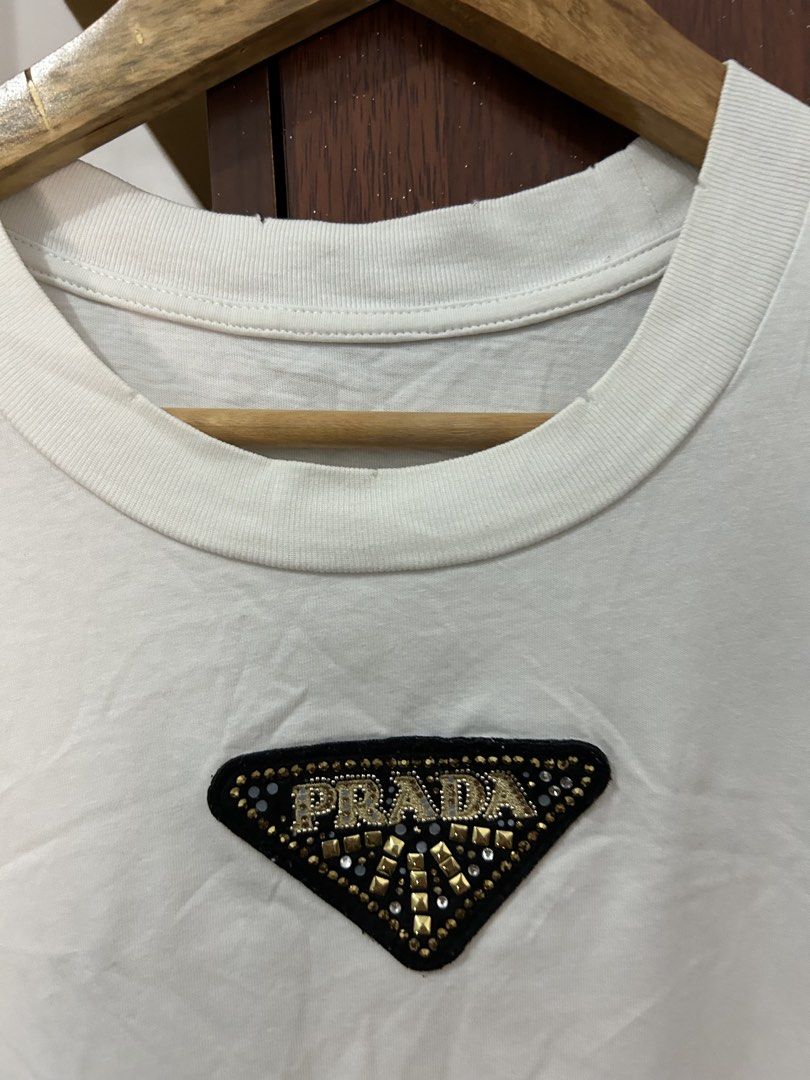 Prada White T-Shirt with Beaded Triangle Logo (Category: Shirts) located in Davao. Submitted by ruggedthrift444 (Listing ID 1465687521, Image 3). Description: - Brand: Prada - Color: White with black triangular chest patch - Details: Embellished gold-tone beads and clear crystals spelling out the brand name on the chest - Fit: Standard relaxed fit - Material: Premium cotton with ribbed collar detailing - Condition: Brand new / Excellent quality -