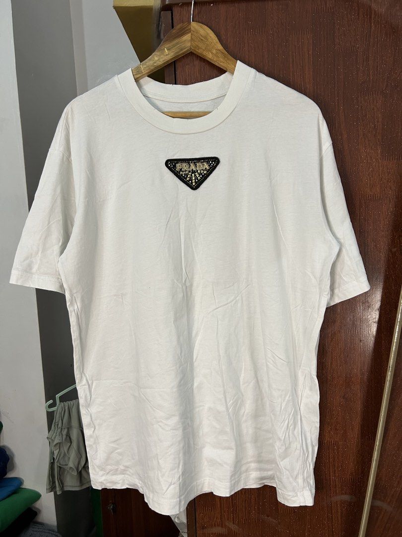 Prada White T-Shirt with Beaded Triangle Logo (Category: Shirts) located in Davao. Submitted by ruggedthrift444 (Listing ID 1465687521, Image 1). Description: - Brand: Prada - Color: White with black triangular chest patch - Details: Embellished gold-tone beads and clear crystals spelling out the brand name on the chest - Fit: Standard relaxed fit - Material: Premium cotton with ribbed collar detailing - Condition: Brand new / Excellent quality -