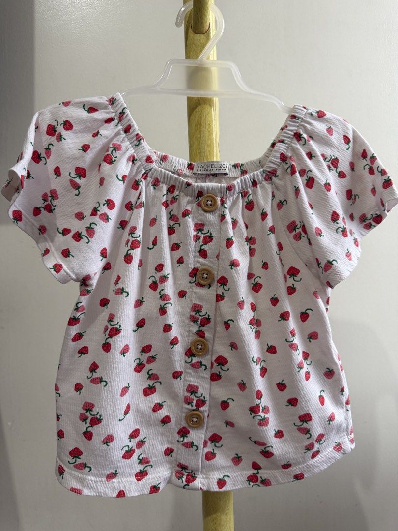 Rachel Zoe White Strawberry Print Button Front Blouse (Category: Babies & Kids Fashion) located in Quezon City. Submitted by warmrabbit_414908 (Listing ID 1465597072, Image 2). Description: - Brand: Rachel Zoe - Design: White fabric with red strawberries and wooden-look front buttons - Style: Cottagecore aesthetic, square collar, short puff sleeves with elastic neckline - Material: Textured fabric - Condition: Excellent pre-loved / as pictured - 