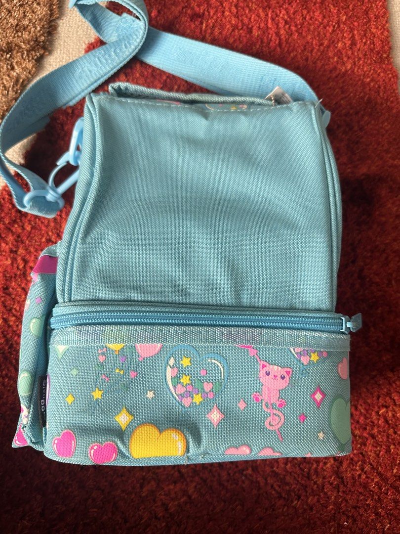 Smiggle light blue heart pattern lunch bag with strap (Category: Babies & Kids Fashion) located in Imus. Submitted by mamibinkz (Listing ID 1465643686, Image 2). Description: - Smiggle double-decker lunch bag in light blue - Adorable all-over print featuring hearts, stars, and little cats - Double-decker zip compartment design for easy food organization - Adjustable shoulder strap with woven Smiggle logo - White Smiggle logo embroidered on the fr