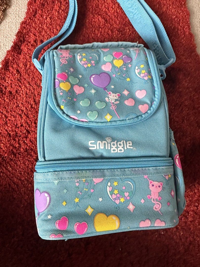 Smiggle light blue heart pattern lunch bag with strap (Category: Babies & Kids Fashion) located in Imus. Submitted by mamibinkz (Listing ID 1465643686, Image 1). Description: - Smiggle double-decker lunch bag in light blue - Adorable all-over print featuring hearts, stars, and little cats - Double-decker zip compartment design for easy food organization - Adjustable shoulder strap with woven Smiggle logo - White Smiggle logo embroidered on the fr