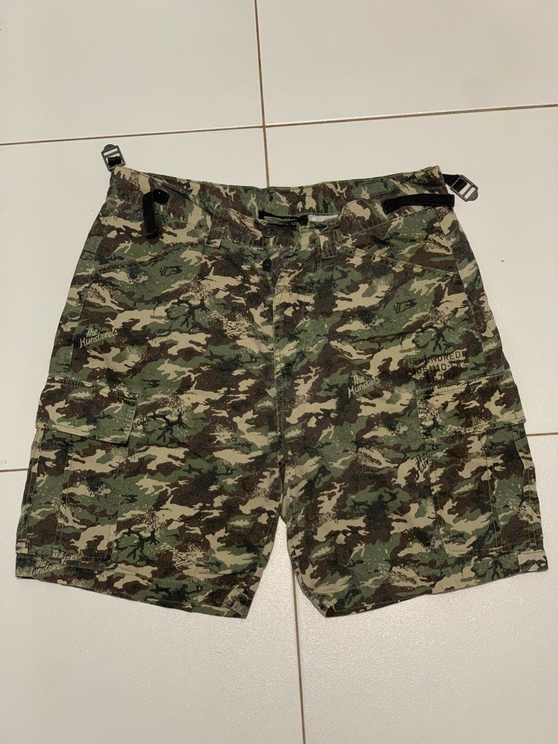 The Hundreds Camo Cargo Shorts (Category: Shorts) located in Mataasnakahoy. Submitted by grailsph.81630 (Listing ID 1465749851, Image 1). Description: - Brand: The Hundreds - Design: All-over camo print (green, brown, black) with subtle script logo accents - Storage: Multiple utility pockets with secure button closures on sides and back - Fit: Adjustable waist straps with metal buckles on both sides - Details: Button fly closure and branded inner
