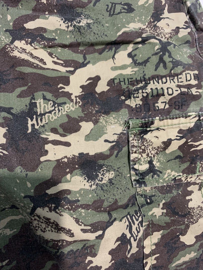 The Hundreds Camo Cargo Shorts (Category: Shorts) located in Mataasnakahoy. Submitted by grailsph.81630 (Listing ID 1465749851, Image 3). Description: - Brand: The Hundreds - Design: All-over camo print (green, brown, black) with subtle script logo accents - Storage: Multiple utility pockets with secure button closures on sides and back - Fit: Adjustable waist straps with metal buckles on both sides - Details: Button fly closure and branded inner