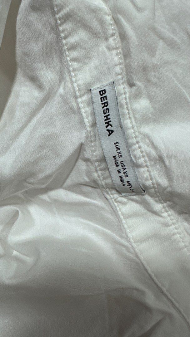 Bershka White Cropped Button-Up Shirt (Category: Shirts) located in San Juan City. Submitted by declutterwithxz (Listing ID 1471178054, Image 2). Description: - Brand: Bershka - Style: Cropped button-down shirt with a relaxed silhouette and dropped shoulders - Details: Pointed collar, single chest pocket, and long sleeves with wide cuffs - Size: XS - Origin: Made in India - Versatility: Essential basic piece, ideal for layering or styling solo wi