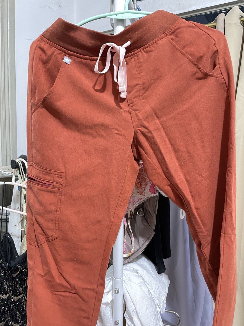 FIGS Rust Orange Scrubs Set Top and Pants (Category: Sets or Coordinates) located in Davao. Submitted by .lille (Listing ID 1471283781, Image 2). Description: FIGS scrubs set in a rust orange shade. V-neck top with chest pocket and branded logo tag. Matching pants with a wide ribbed waistband and adjustable pink drawstring. Multiple pockets including a zippered cargo pocket on the right leg. Technical collection fabric, lightweight and stretchy. 