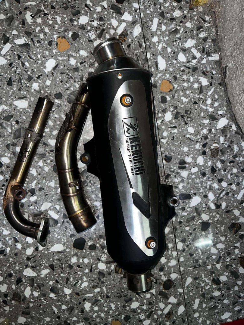 Kenochi Motorcycle Exhaust Pipe System (Category: Mufflers, Exhaust Parts & Accessories) located in Quezon City. Submitted by jcccccccccccccc (Listing ID 1471139904, Image 2). Description: - Brand: Kenochi exhaust pipe setup - Compatibility: Suitable for scooters and underbone motorcycles - Inclusions: Black muffler canister with silver heat shield, header pipes, spring clamps, and mounting points - Condition: Noticeable heavy scratches and wear 