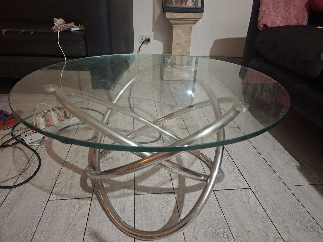 Modern Round Glass Coffee Table with Interlocking Metal Base (Category: Tables & Sets) located in Las Pinas. Submitted by feliboykins (Listing ID 1471214497, Image 1). Description: - Type: Round glass top coffee table - Base: Unique interlocking silver metal ring design - Top: Clear tempered glass secured with metal puck mounts - Condition: Sturdy construction with normal slight everyday wear on the glass surface - Aesthetic: Clean, modern look i