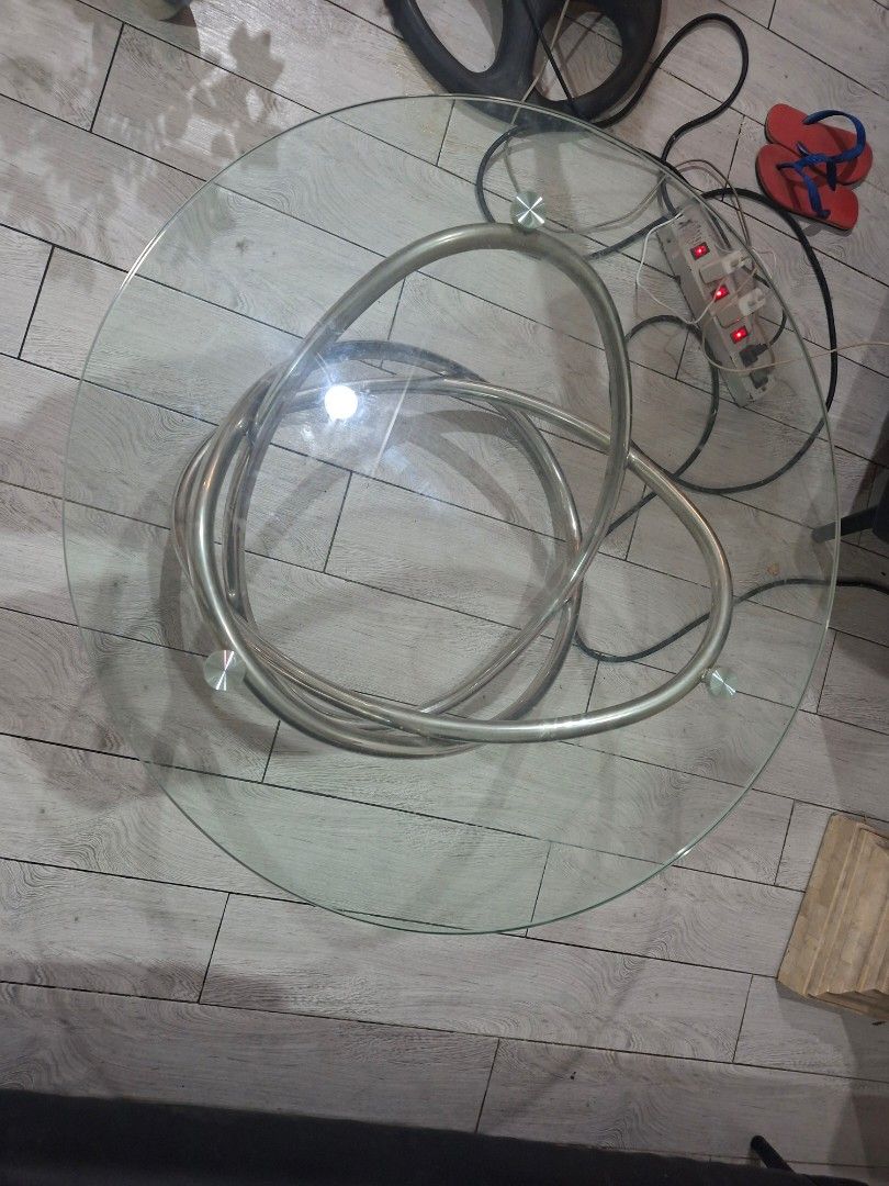 Modern Round Glass Coffee Table with Interlocking Metal Base (Category: Tables & Sets) located in Las Pinas. Submitted by feliboykins (Listing ID 1471214497, Image 3). Description: - Type: Round glass top coffee table - Base: Unique interlocking silver metal ring design - Top: Clear tempered glass secured with metal puck mounts - Condition: Sturdy construction with normal slight everyday wear on the glass surface - Aesthetic: Clean, modern look i