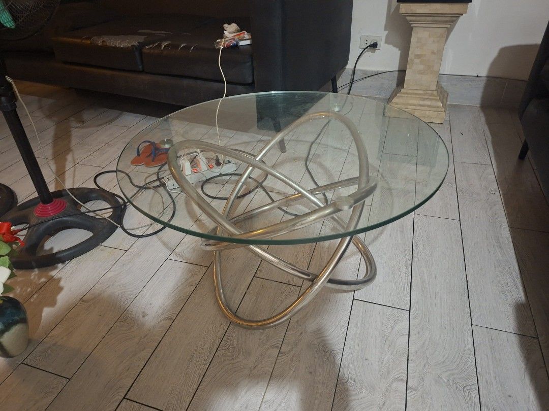 Modern Round Glass Coffee Table with Interlocking Metal Base (Category: Tables & Sets) located in Las Pinas. Submitted by feliboykins (Listing ID 1471214497, Image 2). Description: - Type: Round glass top coffee table - Base: Unique interlocking silver metal ring design - Top: Clear tempered glass secured with metal puck mounts - Condition: Sturdy construction with normal slight everyday wear on the glass surface - Aesthetic: Clean, modern look i