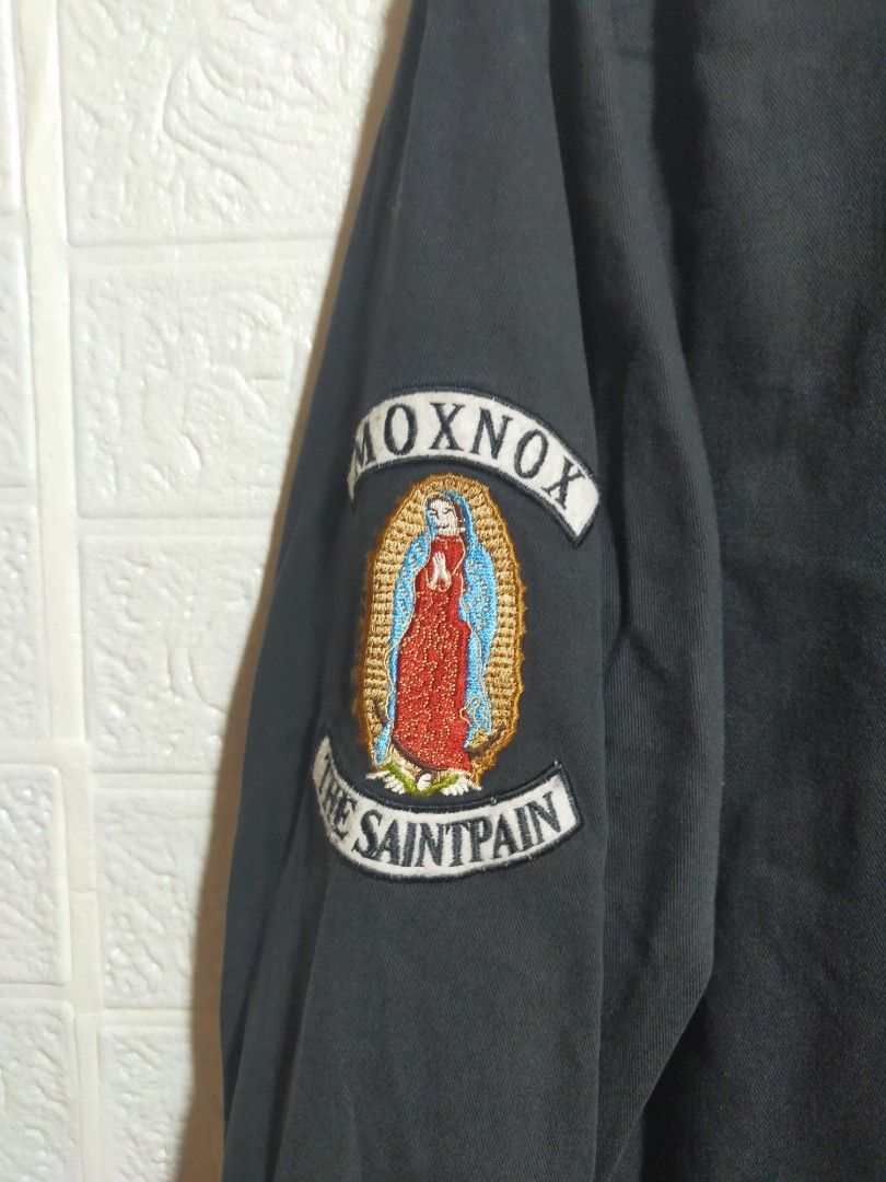 Saintpain Black Bomber Jacket with Patches (Category: Coats, Jackets and Outerwear) located in Valencia. Submitted by cabisaythriftshop (Listing ID 1471223551, Image 2). Description: - Brand: Saintpain - Color: Black - Material: Durable canvas - Closure: Full zip - Details: Ribbed collar, cuffs, and hem - Pockets: Utility pocket with zipper on the left sleeve - Patches: 'Faith & Trust God Bless' chest patch, Virgin Mary right sleeve graphic, and 