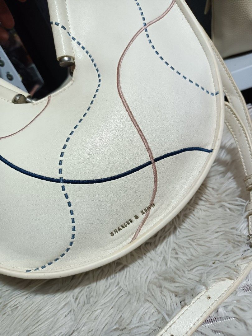 Charles & Keith White Half Moon Bag (Category: Shoulder Bags) located in Naga. Submitted by zksboutique (Listing ID 1465936791, Image 2). Description: - Trendy half moon shape in off-white - Unique blue and pink stitched curved line details on the front - Elegant silver top handles and matching adjustable shoulder strap - Chic gold brand lettering near the bottom edge - Compact and convenient size for daily essentials - Secure zip closure to keep