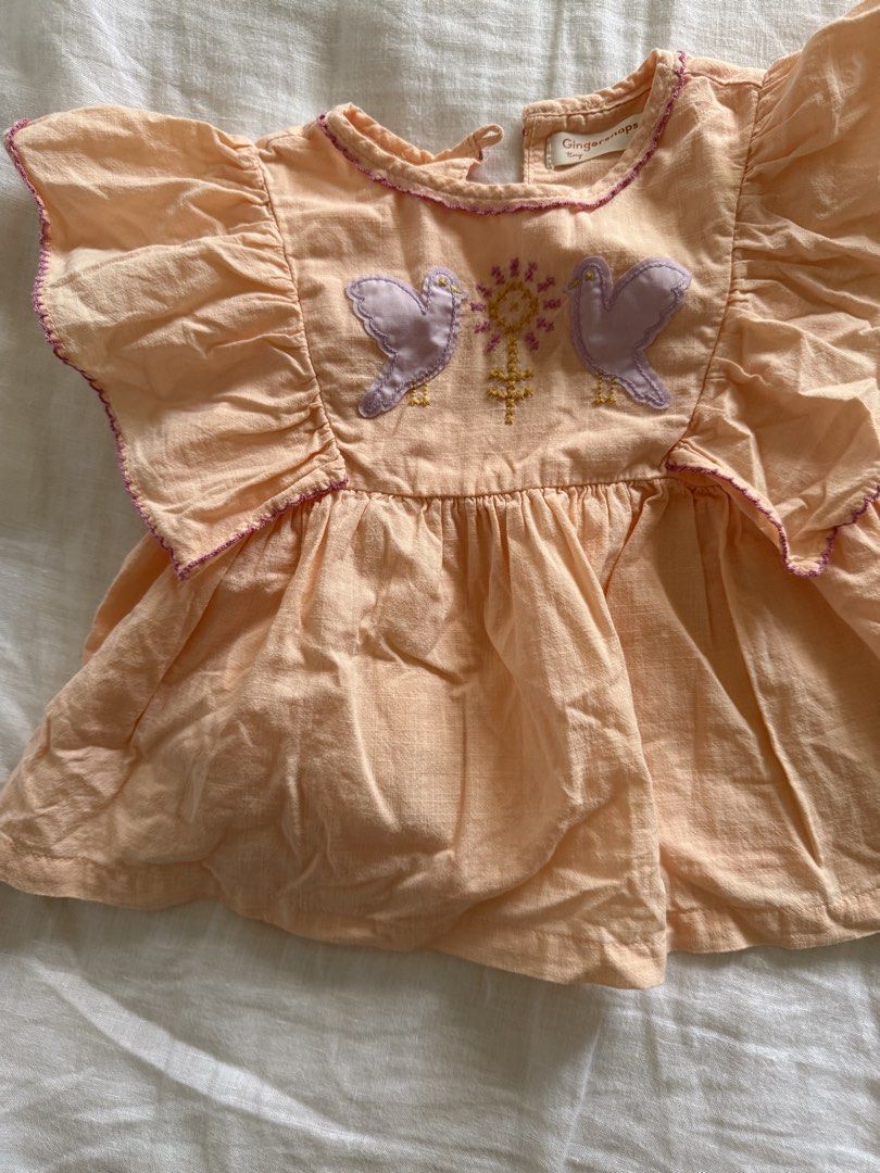 Gingersnaps Tiny Peach Bird Embroidered Flutter Sleeve Top 2Y (Category: Babies & Kids Fashion) located in Taguig. Submitted by muntingkahon (Listing ID 1465765813, Image 2). Description: - Brand: Gingersnaps Tiny - Size: 2Y (fits heights 87-92 cm) - Color: Peach with purple contrast stitching on flutter sleeves and collar - Design: Sweet embroidered purple birds and floral details on the front - Closure: Convenient button closure at the back nec