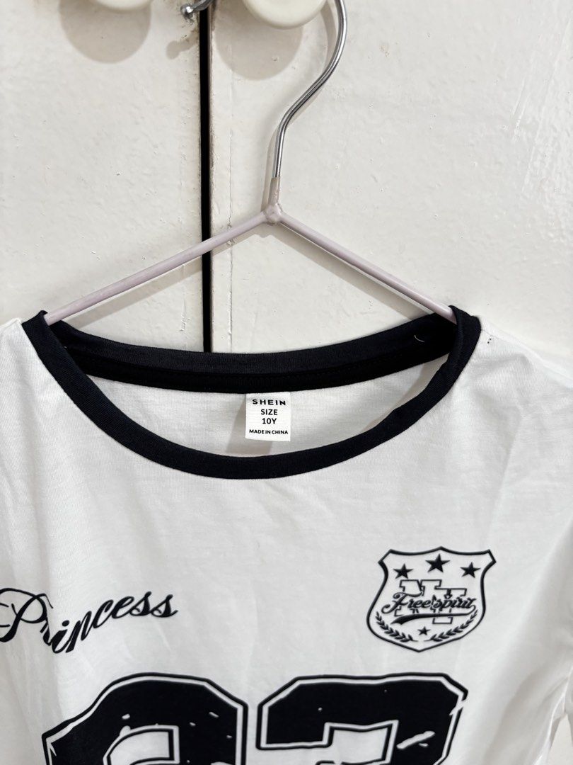 SHEIN White Graphic Ringer Shirt (Category: Shirts) located in Manila. Submitted by dianafayesimbre (Listing ID 1465831209, Image 3). Description: - Brand/Size: SHEIN Kids Size 10Y on tag but can fit small to semi large on womens - Design: White ringer tee with black collar trim, large '23' print, 'Princess' script, and crest logo - Accents: Cute black heart graphics on both sleeves - Material: 100% cotton, relaxed fit for everyday comfort - Cond