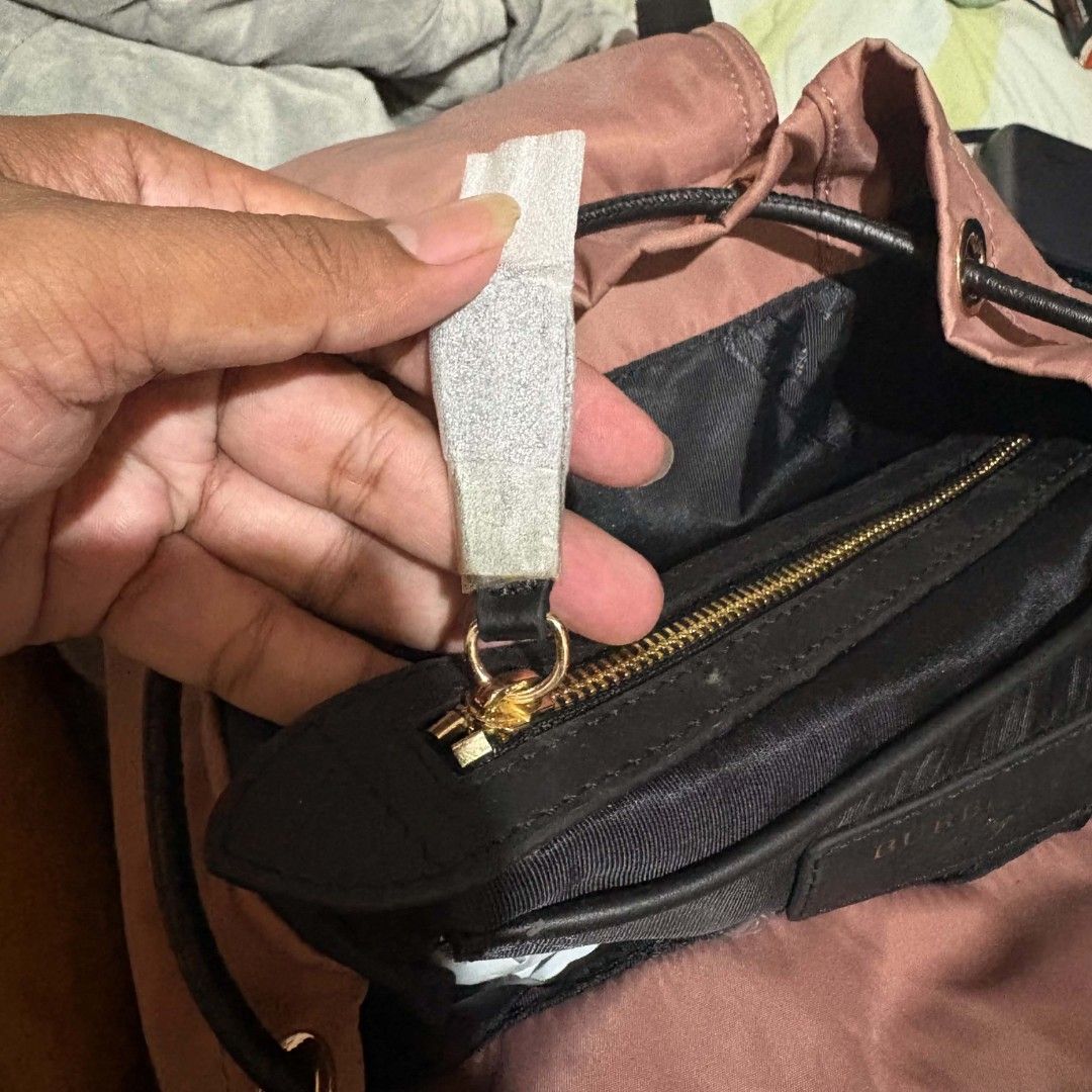 Burberry The Rucksack Cloth Backpack Dusty Pink (Category: Backpacks) located in Carmen. Submitted by ar_thrift00 (Listing ID 1466088608, Image 3). Description: - Brand new with original tags attached - Color/Material: Dusty pink nylon cloth with black leather trim - Hardware: Elegant gold-tone accents - Closure: Secure drawstring with top buckle flap - Pockets: Front zip pocket, two side zip pockets, and interior zip pocket - Interior: Features 