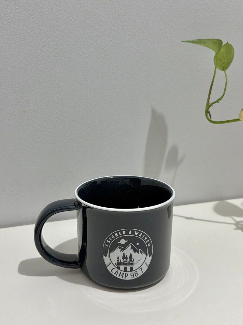Camp 98.7 Mountain Black Ceramic Mug (Category: Coffee & Tea Tableware) located in Makati City. Submitted by ariannelaz54133 (Listing ID 1466154807, Image 2). Description: - Material: Black ceramic with a glossy finish and white inner rim - Design: "Camp 98.7" text printed on one side - Graphic: Round mountain graphic with "I Signed A Waiver" and "Camp 98.7" on the reverse side - Handle: Standard side handle for a comfortable grip - Condition: Br