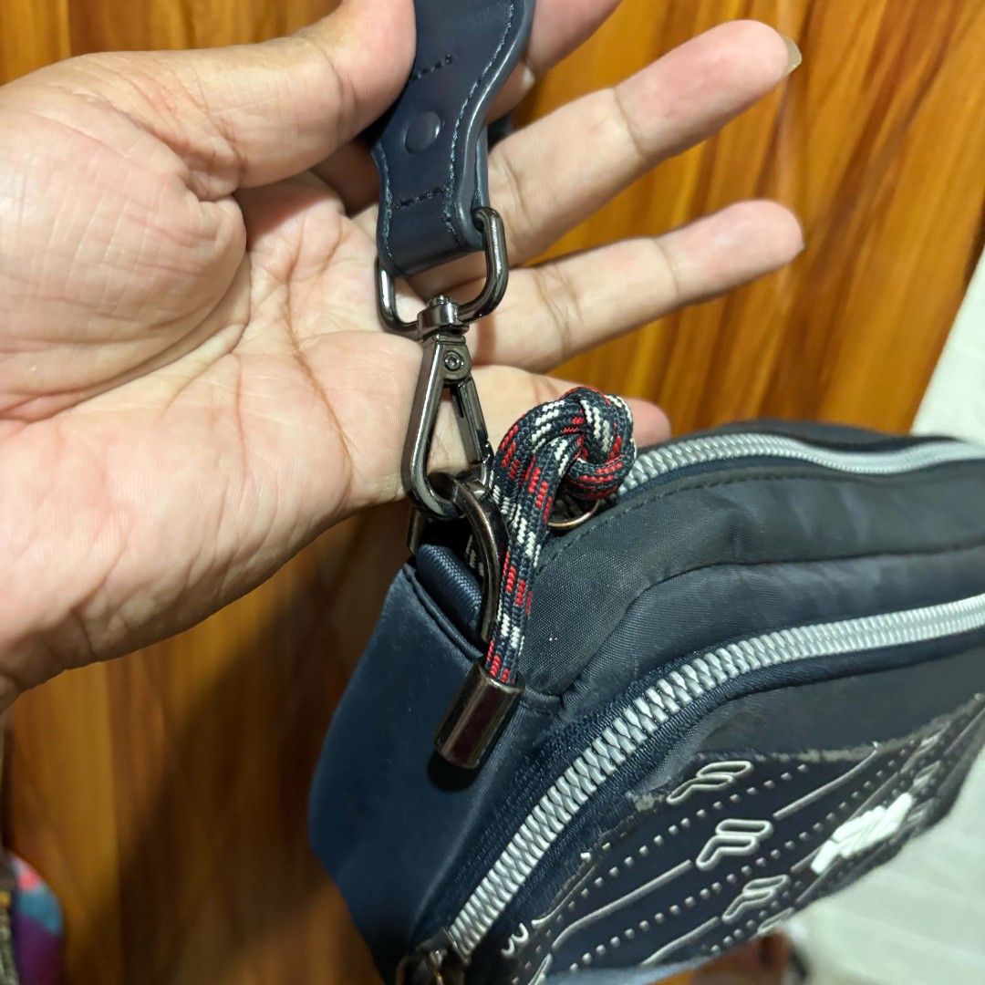 Fila Crossbody Bag (Category: Cross-body Bags) located in Carmen. Submitted by ar_thrift00 (Listing ID 1466085108, Image 3). Description: - Brand: Fila - Color: Navy blue with white logo prints and diagonal stripe patterns - Strap: Adjustable with metal eyelets and side clips - Interior: Fabric lining with brand tags and serial number labels - Closure: Zipper closures with braided red, white, and black pull cords - Condition: Minor wear near the 