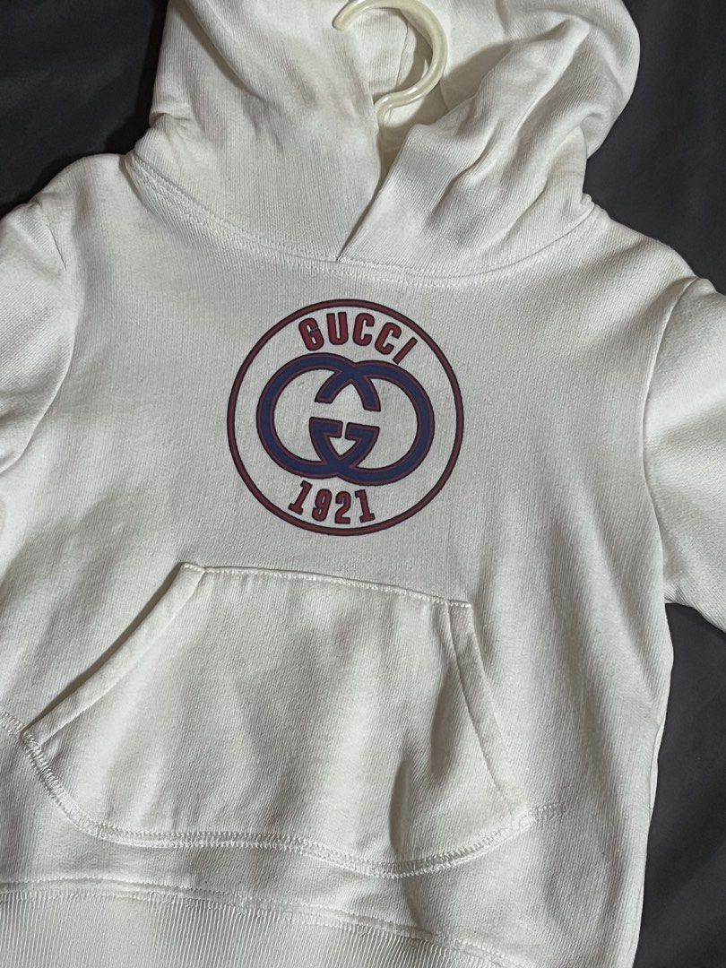 Gucci 1921 Logo White Hoodie (Authentic) (Category: Babies & Kids Fashion) located in Cavite. Submitted by mariellesophia (Listing ID 1465993425, Image 1). Description: - Authentic white Gucci hoodie with the iconic 1921 circular chest graphic - Classic pullover design featuring an attached hood and front pouch pocket - Clean fabric with ribbed cuffs and hem that hold their shape well - Soft interior lining for maximum warmth and everyday comfort