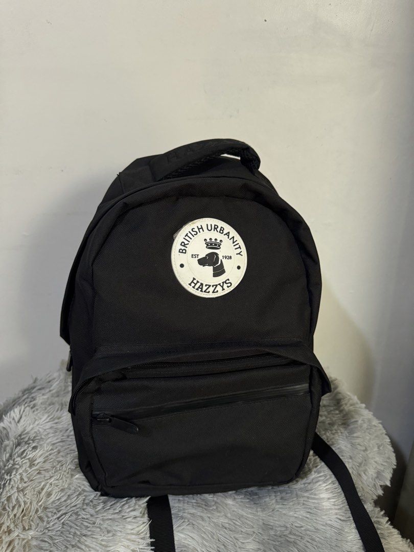HAZZYS British Urbanity Black Backpack (Category: Backpacks) located in Urdaneta. Submitted by kathleenja24136 (Listing ID 1465957738, Image 2). Description: - Brand: HAZZYS - Material: Durable black canvas - Design: Large circular logo patch with crown & dog graphic and 'BRITISH URBANITY' text - Details: 'HAZZYS' embroidered grab handle and front zippered pocket - Comfort: Adjustable shoulder straps - Use: Ideal everyday carry for school or comm