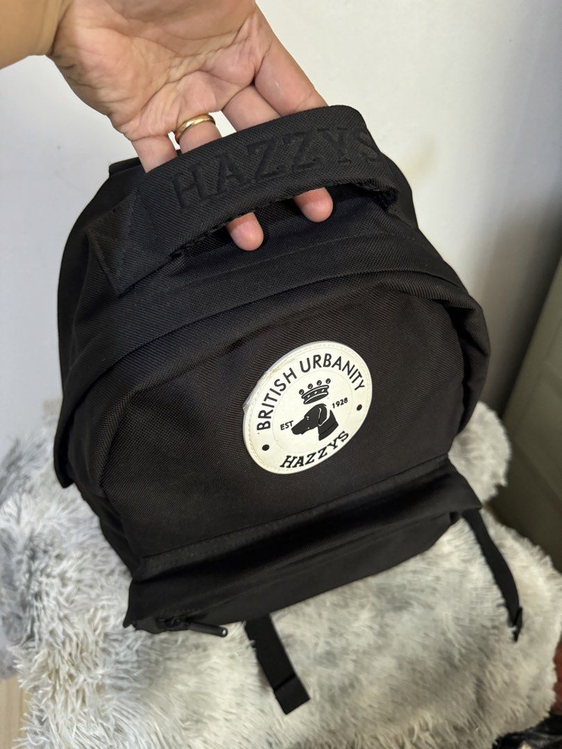 HAZZYS British Urbanity Black Backpack (Category: Backpacks) located in Urdaneta. Submitted by kathleenja24136 (Listing ID 1465957738, Image 3). Description: - Brand: HAZZYS - Material: Durable black canvas - Design: Large circular logo patch with crown & dog graphic and 'BRITISH URBANITY' text - Details: 'HAZZYS' embroidered grab handle and front zippered pocket - Comfort: Adjustable shoulder straps - Use: Ideal everyday carry for school or comm