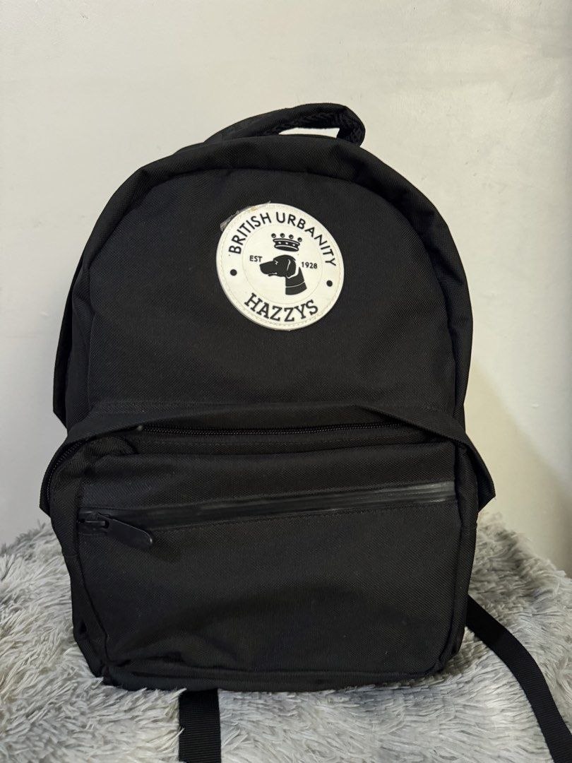 HAZZYS British Urbanity Black Backpack (Category: Backpacks) located in Urdaneta. Submitted by kathleenja24136 (Listing ID 1465957738, Image 1). Description: - Brand: HAZZYS - Material: Durable black canvas - Design: Large circular logo patch with crown & dog graphic and 'BRITISH URBANITY' text - Details: 'HAZZYS' embroidered grab handle and front zippered pocket - Comfort: Adjustable shoulder straps - Use: Ideal everyday carry for school or comm