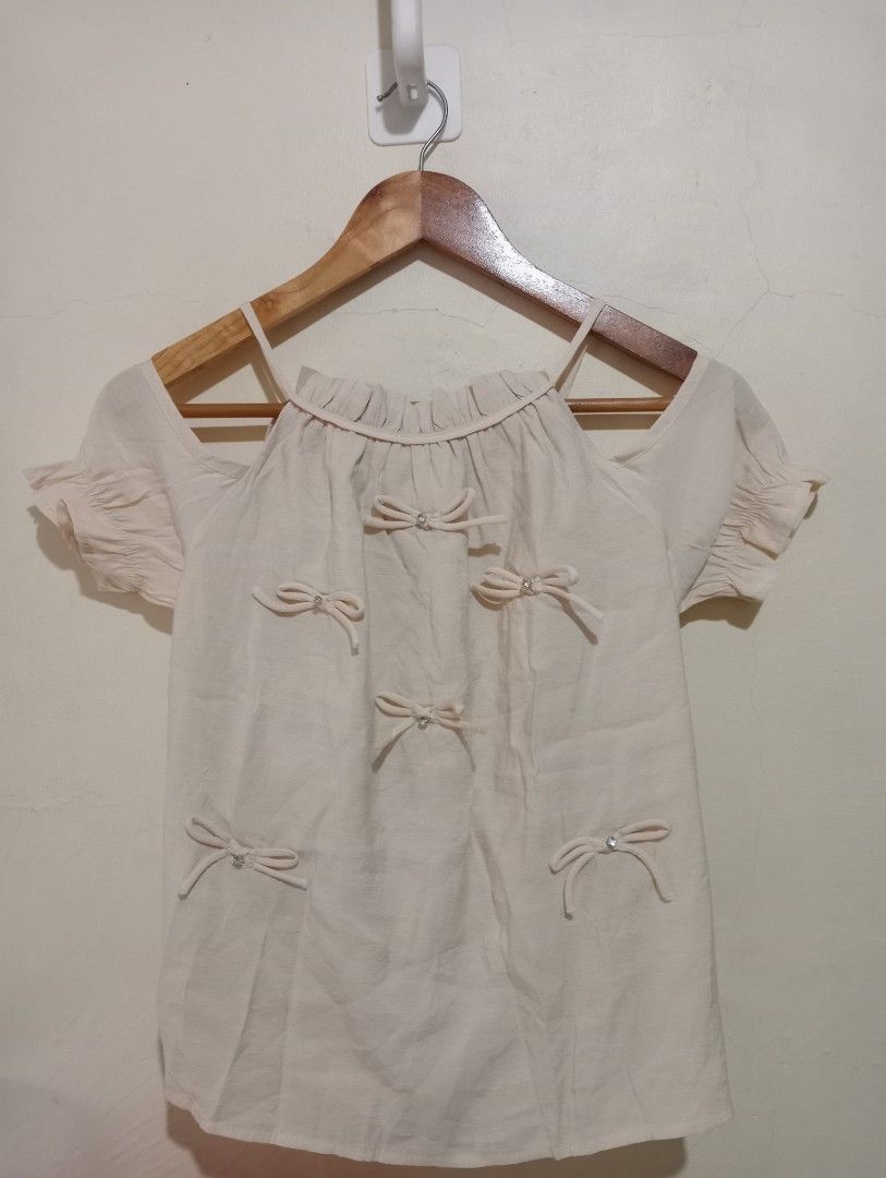 Off-shoulder beige blouse with bow details (Category: Blouses) located in Manila. Submitted by purplethings28 (Listing ID 1466006705, Image 2). Description: - Color: Beige - Style: Off-shoulder top with short sleeves and ruffled neckline - Details: Cute bow appliques with tiny crystal rhinestones in the center - Fabric: Lightweight and comfortable - Condition: Clean, excellent, and ready to wear - Styling tip: Great for pairing with high-waisted 