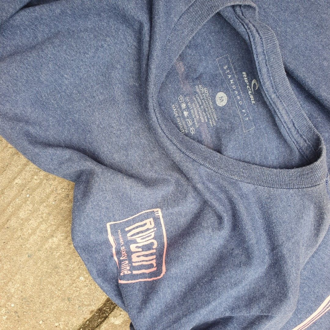 Rip Curl 1969 Graphic Long Sleeve Blue Medium (Category: Tshirts & Polo Shirts ) located in Cavite. Submitted by josephnl (Listing ID 1466130500, Image 3). Description: - Brand: Rip Curl - Style: Long sleeve shirt with standard fit - Color: Heather blue with pink accents - Design: Large pink graphic on back, smaller logo on front neck, and vertical pink stripes on sleeve - Size: Medium - Material: Cotton and polyester blend - Measurements: Width 