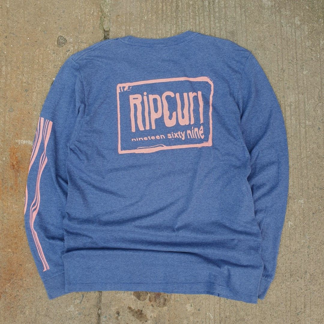 Rip Curl 1969 Graphic Long Sleeve Blue Medium (Category: Tshirts & Polo Shirts ) located in Cavite. Submitted by josephnl (Listing ID 1466130500, Image 1). Description: - Brand: Rip Curl - Style: Long sleeve shirt with standard fit - Color: Heather blue with pink accents - Design: Large pink graphic on back, smaller logo on front neck, and vertical pink stripes on sleeve - Size: Medium - Material: Cotton and polyester blend - Measurements: Width 