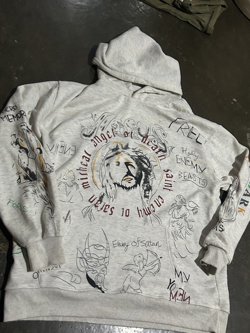 Saint Michael Jesus Print Hoodie (Category: Hoodies) located in Cabuyao City. Submitted by hachikuro (Listing ID 1466039799, Image 2). Description: - Design: Grey pullover hoodie with all-over hand-drawn style illustrations and graphic text - Back Details: Large 'SAINT' lettering paired with an angel graphic 25.5 x27 issue one penhole naka post na po and no tags.