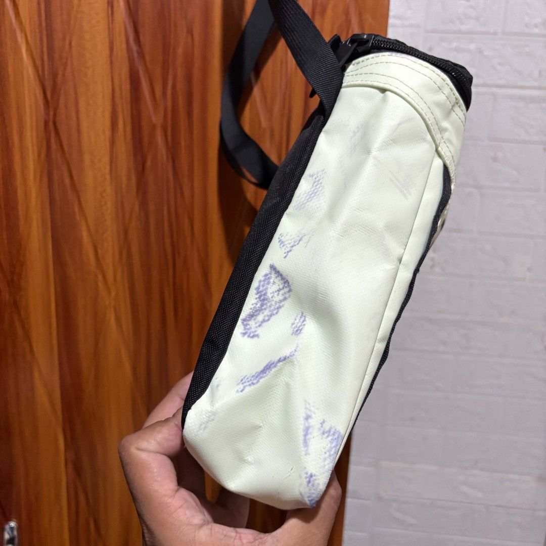 The North Face Crossbody Bag (Category: Sling Bags) located in Carmen. Submitted by ar_thrift00 (Listing ID 1466084642, Image 3). Description: - Brand: The North Face - Color: Off-white with bold black front logo - Features: Adjustable black strap, top zipper closure, vertical front daisy chain webbing - Condition: Good overall, but has minor colored ink/dye stains on the side and back panels (fabric otherwise clean) - Perfect for casual streetwe