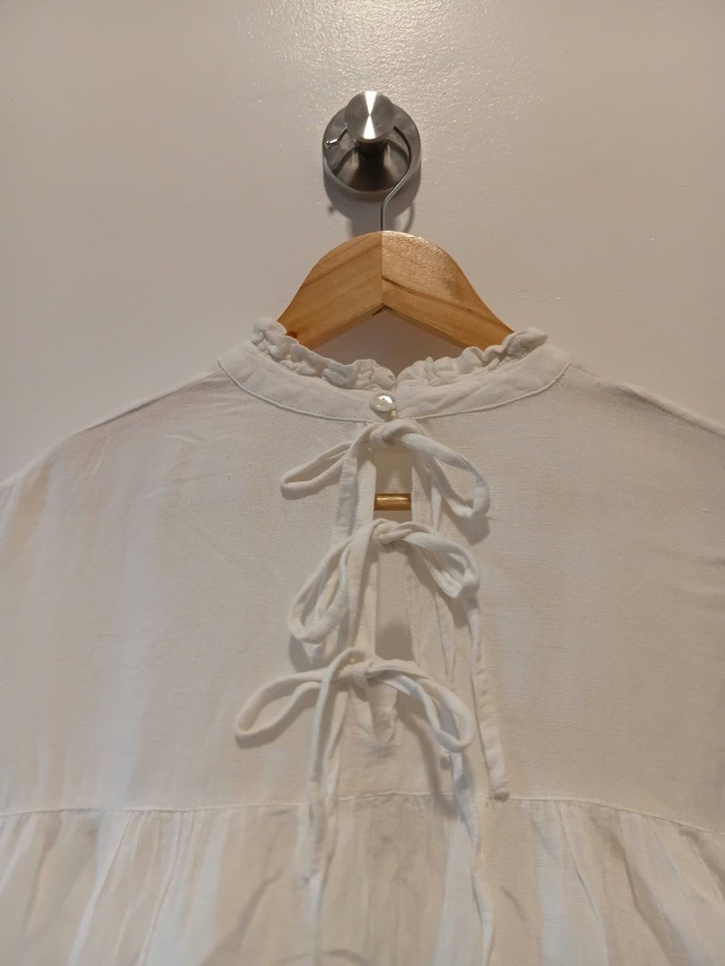 White ruffle neck tie back blouse M-L (Category: Blouses) located in Antipolo. Submitted by wisecat.4a7eea (Listing ID 1466040489, Image 3). Description: - Color: White - Size: Fits Medium to Large - Design: High ruffled neckline with single back button - Details: Cute back cutout with three ribbon ties and gathered front/back yoke - Sleeves: Mid-length with cuffs - Fabric: Lightweight and comfortable.