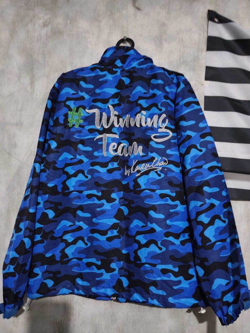 Blue Camo Winning Team Windbreaker Jacket (Category: Coats, Jackets and Outerwear) located in Baguio. Submitted by fordaluvofthrift2600 (Listing ID 1466168556, Image 2). Description: - Color/Pattern: Blue and black camo print - Closures & Pockets: White front zipper and functional side zip pockets - Hem: Adjustable drawstring for a customized fit - Back Detail: Embroidered 'Winning Team' lettering in silver with a green hash symbol and signature 