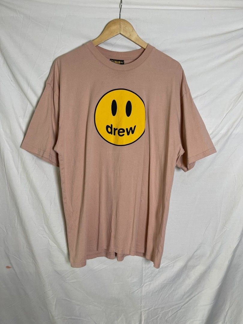 drew house mascot tee pink size L (Category: Shirts) located in Manila. Submitted by japee55 (Listing ID 1466320939, Image 1). Description: Drew house mascot t-shirt in pink. Large yellow smiley face print on the front with black 'drew' text. Size tag says L. Drew house neck tag and care label attached inside. Small stain dots on the lower side fabric as pictured. Comfortable cotton everyday tee..