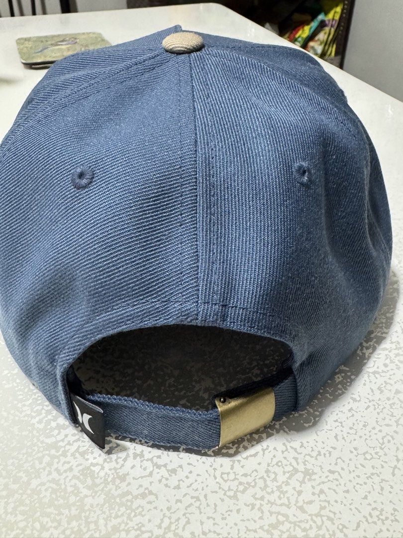 Hurley The Classics Yupoong Snapback Cap (Category: Caps & Hats) located in Cavite. Submitted by marcuz1316 (Listing ID 1466255066, Image 2). Description: Hurley hat by Yupoong, made in Bangladesh. Blue crown with khaki visor and top button. Front has a woven patch with palm trees, sunset, and a camper van graphic. Adjustable metal clasp strap on the back with small Hurley logo tag. Interior has The Classics Yupoong branding tag, 80 percent acryl