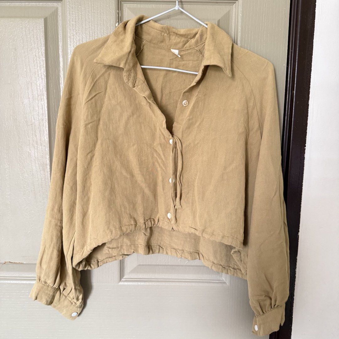 Khaki beige cropped button-down shirt blouse long sleeves (Category: Longsleeves) located in Angeles City. Submitted by elliemnopqrstuv (Listing ID 1466206713, Image 2). Description: - Color: Khaki beige - Style: Cropped button-down shirt with a loose, relaxed fit - Details: Collared neckline, white front buttons, long sleeves with gathered cuffs, and a tie string hem - Fabric: Soft, textured material - Styling: Great to pair with high-waisted je