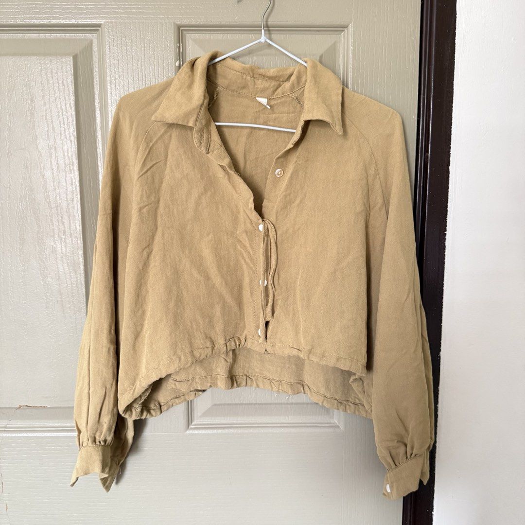 Khaki beige cropped button-down shirt blouse long sleeves (Category: Longsleeves) located in Angeles City. Submitted by elliemnopqrstuv (Listing ID 1466206713, Image 1). Description: - Color: Khaki beige - Style: Cropped button-down shirt with a loose, relaxed fit - Details: Collared neckline, white front buttons, long sleeves with gathered cuffs, and a tie string hem - Fabric: Soft, textured material - Styling: Great to pair with high-waisted je