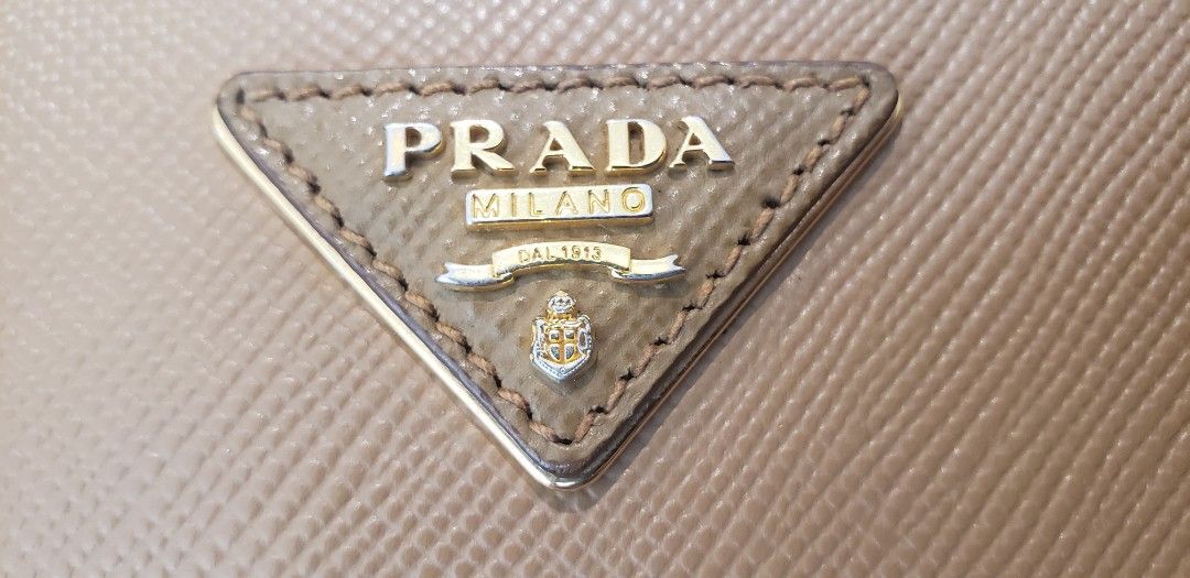 Prada Saffiano Leather Tote Bag (Category: Luxury Bags & Wallets) located in Marikina City. Submitted by racoon143 (Listing ID 1466200454, Image 2). Description: Prada tote bag in light brown Saffiano leather. Gold hardware and metal lettering logo on the front triangular plaque. Double top handles. Red leather interior lining with Prada heatstamp branding. Includes matching leather luggage tag with gold Prada lettering. Clean structure, firm sha