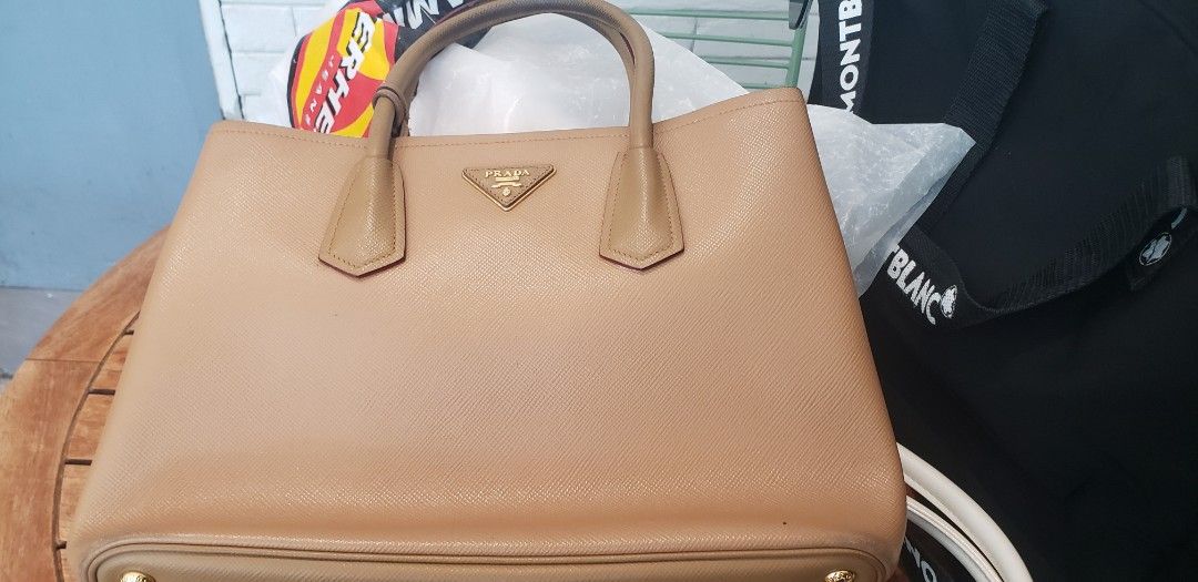 Prada Saffiano Leather Tote Bag (Category: Luxury Bags & Wallets) located in Marikina City. Submitted by racoon143 (Listing ID 1466200454, Image 1). Description: Prada tote bag in light brown Saffiano leather. Gold hardware and metal lettering logo on the front triangular plaque. Double top handles. Red leather interior lining with Prada heatstamp branding. Includes matching leather luggage tag with gold Prada lettering. Clean structure, firm sha