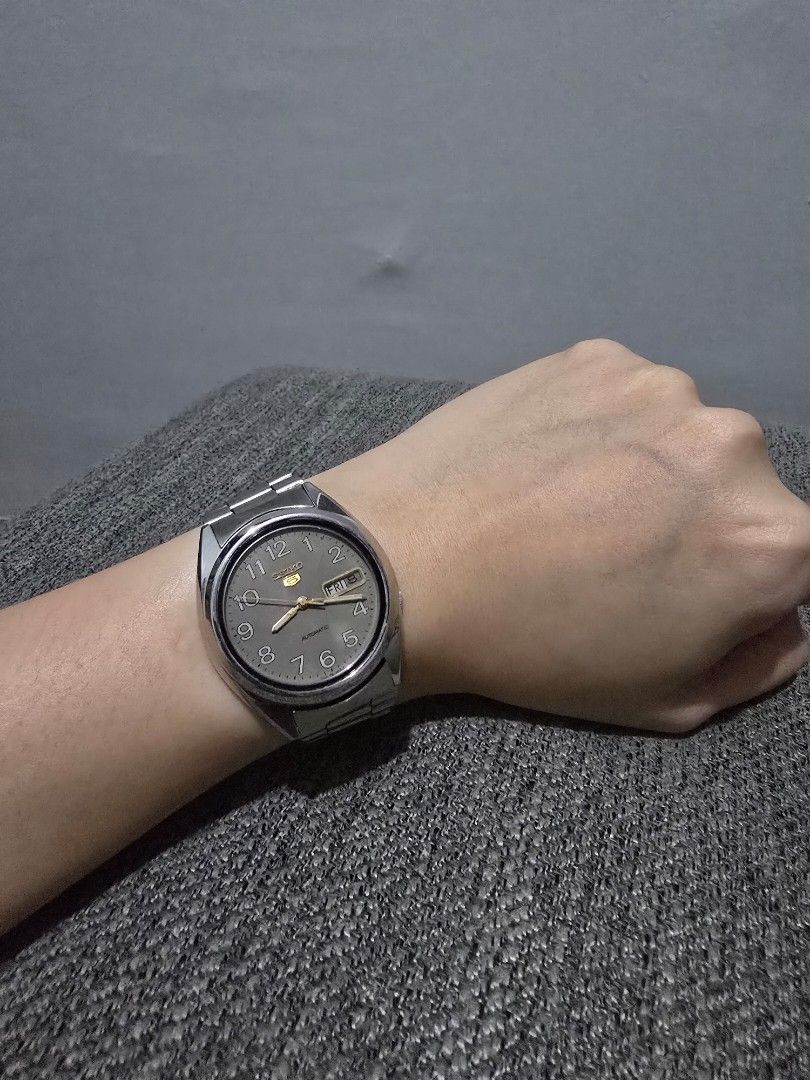 Seiko 5 Automatic Watch with Grey Arabic Numeral Dial (Category: Men's Watches) located in Manila. Submitted by dunnohihi (Listing ID 1466269297, Image 1). Description: Seiko 5 automatic watch on wrist. Stainless steel case and matching metal bracelet. Grey sunburst dial featuring Arabic numerals around the outer edge. Day and date display positioned at 3 o'clock. Gold-tone hands and hour markers. Signed Seiko clasp on the band. Classic everyday 