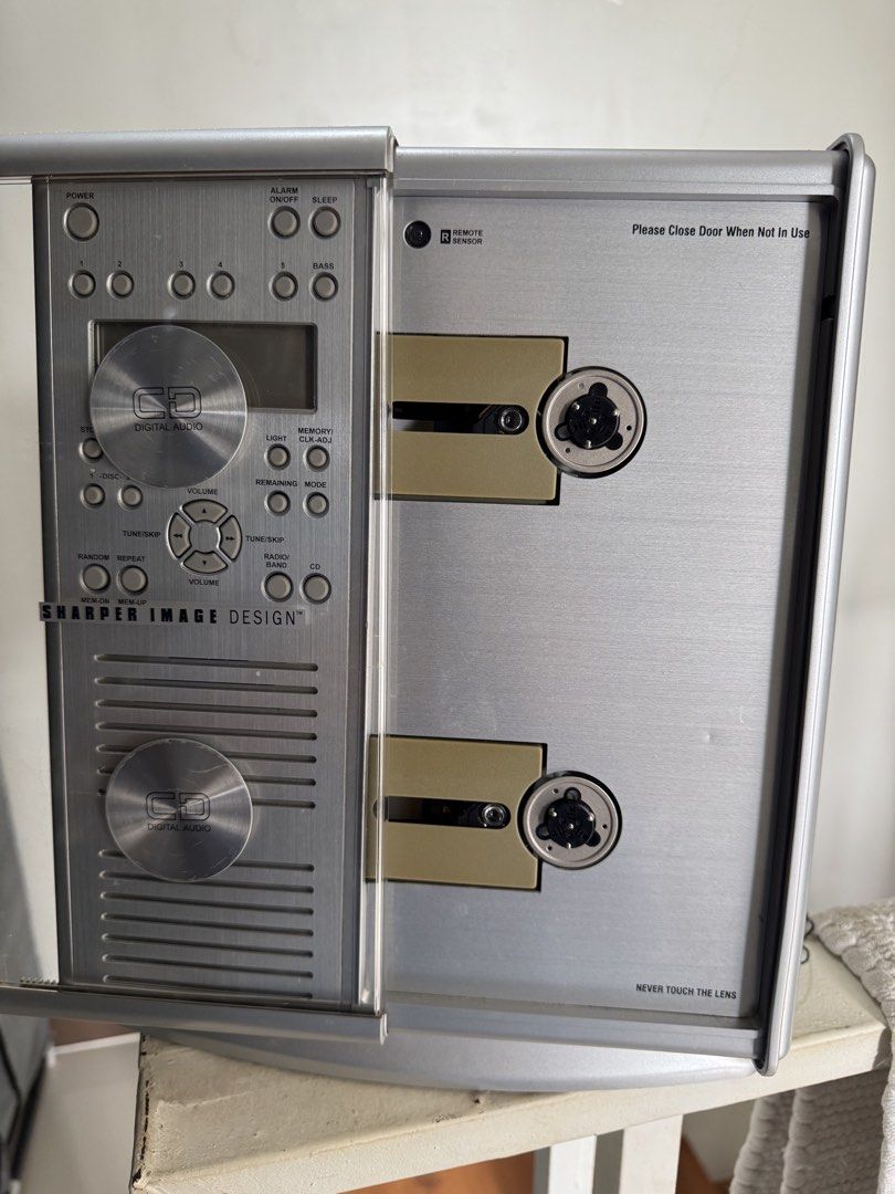 Sharper Image Design CD Digital Audio Player (Category: Portable Music Players) located in Cavite. Submitted by marcuz1316 (Listing ID 1466254404, Image 2). Description: - Brand: Sharper Image Design - Color: Sleek silver finish with round metallic door covers - Design: Vertical dual CD slot setup with a central digital display - Controls: Left-side panel for power, alarm, volume, and playback - Inclusions: Comes with the original remote control 