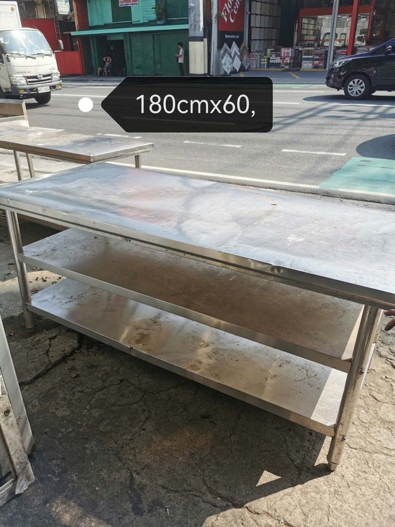 Stainless Steel Kitchen Work Table with 2 Shelves (Category: Tables & Sets) located in Manila. Submitted by marquezpa52205 (Listing ID 1466212692, Image 1). Description: - Heavy-duty stainless steel kitchen/restaurant prep table - Dimensions: 180cm x 60cm - Features two lower storage shelves and sturdy metal legs - Condition: Shows significant rust spots and stains from previous use - Needs a good scrub and polish to restore its shine - Grab a ba