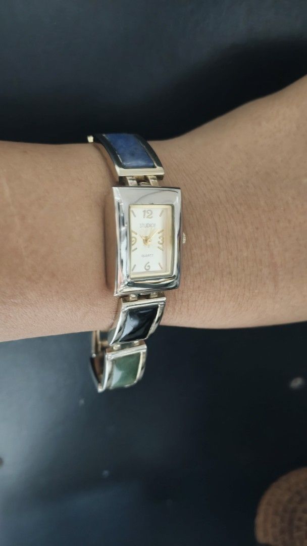 Studio D Quartz Watch with Multicolored Stone Bracelet (Category: Women's Watches) located in Taguig. Submitted by mercyeugen28914 (Listing ID 1466287985, Image 3). Description: - Brand: Studio D - Movement: Battery-operated quartz - Dial: Rectangular face with gold-tone Arabic numerals at 12, 3, and 6 - Strap: Bracelet link design featuring vibrant multicolored rectangular stones (blue, green, red, yellow, black) - Casing: Silver-tone metal with